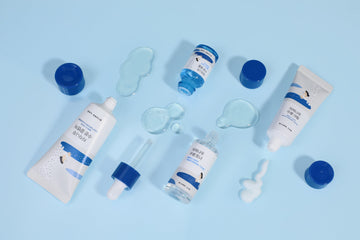 Korean skincare tubes, bottles, and serum with blue caps on light blue background, product textures visible