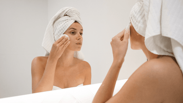 Woman in towel applying skincare with cotton pad in bathroom mirror