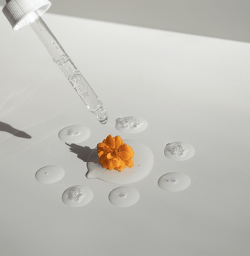 Skincare serum dropper hovering over cream and an orange flower on a white surface