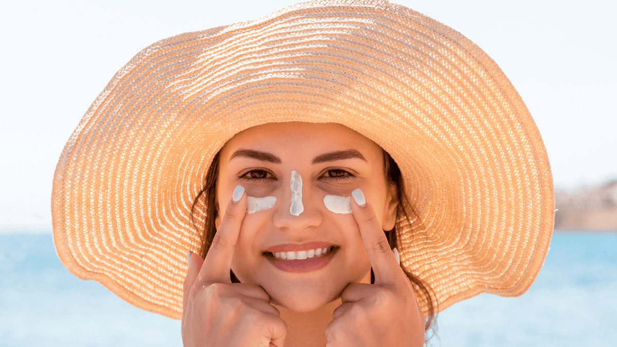 Woman applying sunscreen to her cheeks and nose while wearing a wide-brimmed hat at the beach