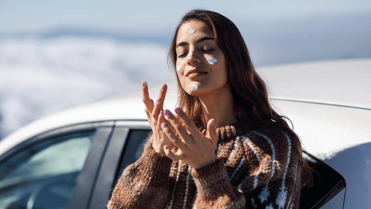 Woman applying sunscreen to her face beside a car in a snowy winter setting