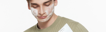 Man with white cleanser foam on his face wearing a green sweater against a white background