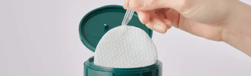 Hand lifting a cotton pad from an open teal skincare container