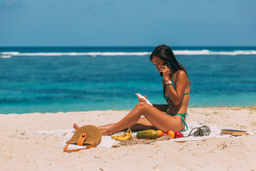 Woman sitting on a beach holding a skincare bottle while talking on her phone