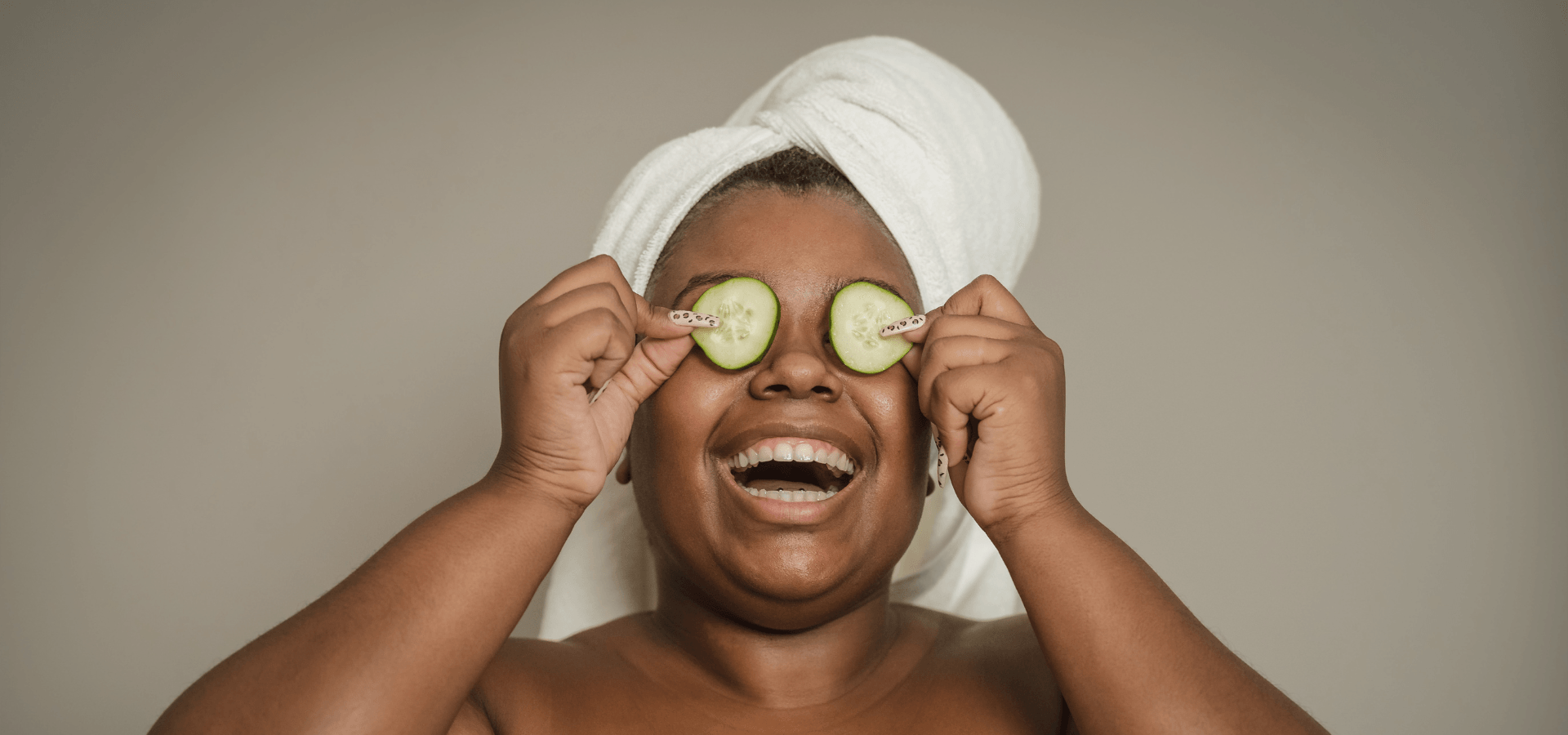 Smiling woman with towel on head holding cucumber slices over eyes, skincare concept