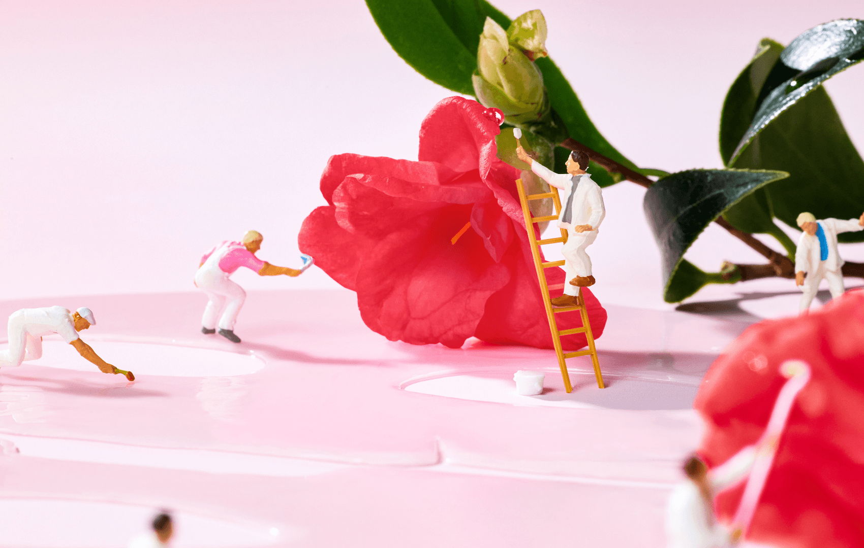 Miniature people painting around a pink camellia flower with glossy petals and green leaves