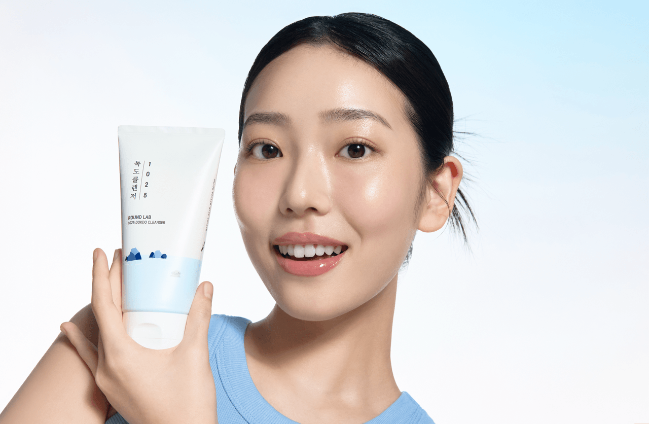 Smiling woman with glowing skin holds 1025 Dokdo Cleanser against a light blue background