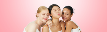 Three women with glowing, clear skin posing together on a pink background, skincare concept