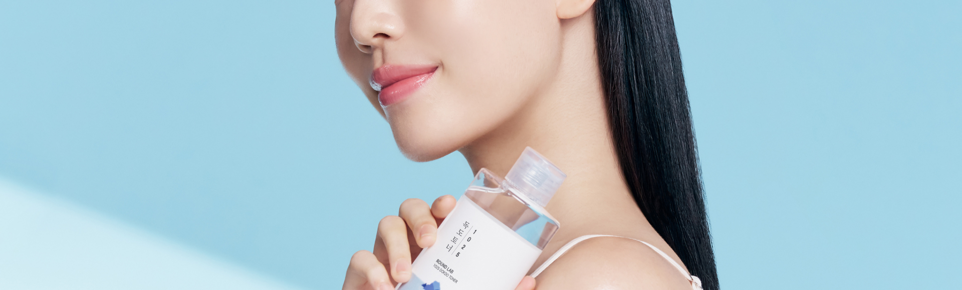 Woman with clear skin holding toner bottle, promoting Korean clean skincare on blue background