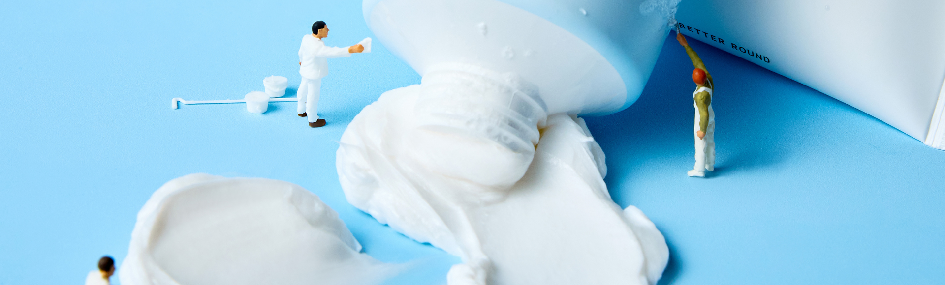 Miniature figures examining spilled skincare cream from a tube on a blue background