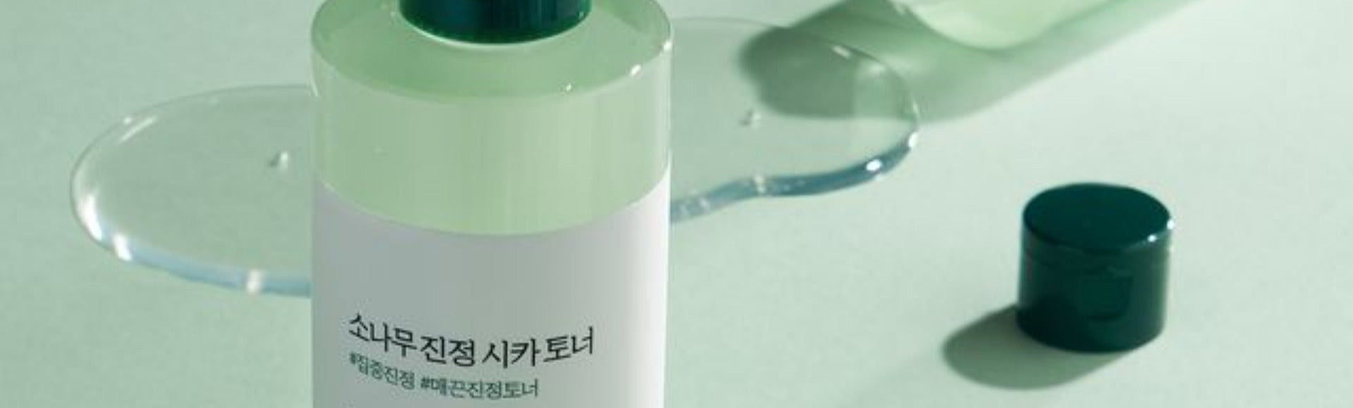 Korean skincare toner bottle with green cap and spilled liquid on pastel surface