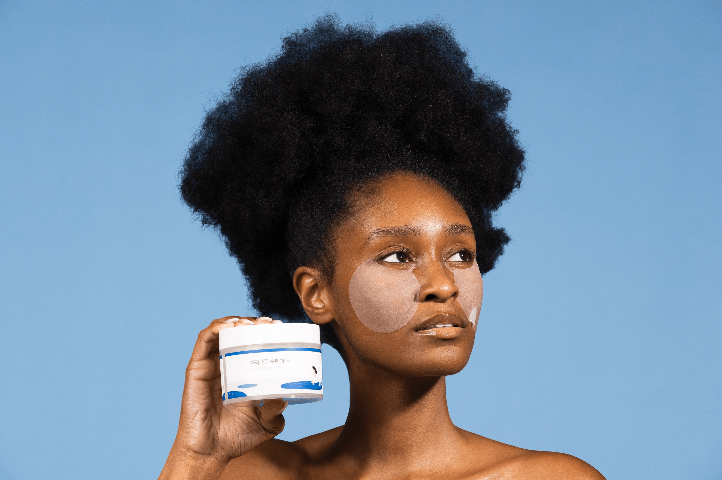 Woman with under-eye hydrogel patches holding a Korean skincare pad container against a blue background.